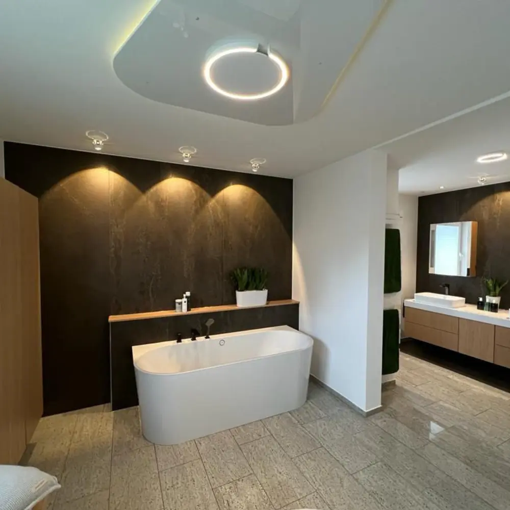 Elegantes Badezimmer mit freistehender Badewanne vor einer dunklen XXL-Keramik-Wandverkleidung in Natursteinoptik, warmem Akzentlicht, Eichenholz-Waschtisch und hellem Steinboden – realisiert in Korschenbroich bei Düsseldorf<br />
